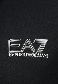 Μαύρο ύφασμα με λευκό στρωματοποιημένο κείμενο που γράφει "EA7 EMPORIO ARMANI" και το λογότυπο του αετού Armani στο κέντρο.