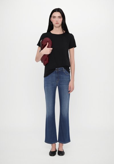 Sort t-shirt med korteærmer og rund halsudskæring, parret med blå flagrende jeans. Modellen holder en bordeaux taske og har sorte ballerinaer på.