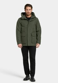 Mies, jolla on yllään oliivinvihreä huppari, edessä taskut, mustat housut ja mustat saappaat, seisoo yksinkertaista vaaleaa taustaa vasten.