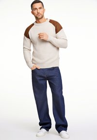 Beige geripptes Sweatshirt mit braunen Wildleder-Schulterakzenten, kombiniert mit dunkelblauen weit geschnittenen Jeans und weißen Turnschuhen.
