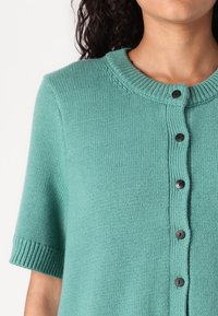 Short-sleeve cardigan in soft, textured teal knit, featuring a round neckline and black button front closure. Ribbed sleeve cuffs.