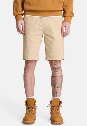 Uomo con pantaloncini beige, stivali marroni, felpa color senape; tatuaggio sulla gamba destra mostra una faccina sorridente, ali, spada e testo.