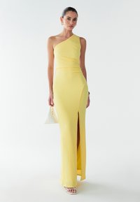 Asymmetrisches gelbes Kleid mit strukturierter Oberfläche, das einen seitlichen Schlitz aufweist; kombiniert mit einer weißen Handtasche und offenen weißen Sandalen.