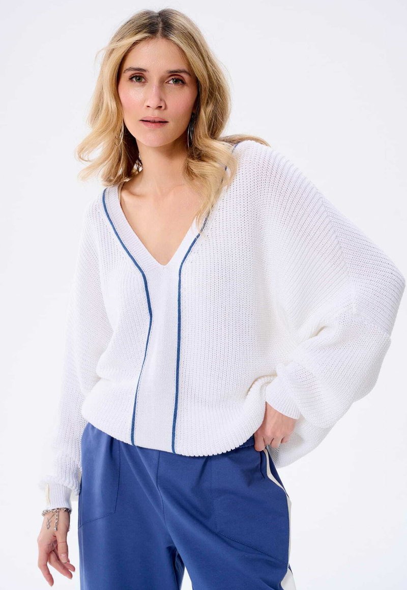 Witte gebreide trui met V-hals, blauwe accenten en een losse fit, gecombineerd met blauwe broek. De trui heeft een ontspannen ontwerp en een gestructureerde stof.