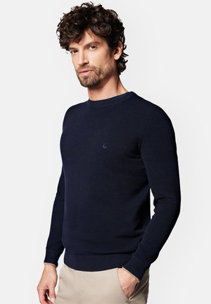 Granatowy sweter dzianinowy z okrągłym dekoltem, ściągaczami przy rękawach i u dołu, z małym logo na piersi. Gładka tekstura, dopasowany krój.