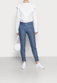 Blusa bianca a maniche lunghe con colletto arricciato e texture a motivi, abbinata a pantaloni aderenti azzurri e scarpe da ginnastica bianche, che tiene una piccola borsa bianca.