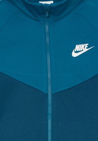 Casaco com fecho éclair em teal com colarinho alto, apresentando o logo branco da Nike no canto superior esquerdo. Textura suave, design em bloco de cores contrastantes.
