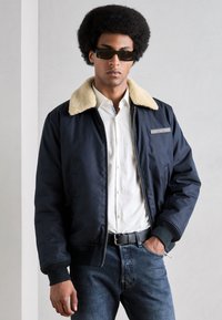 Chaqueta bomber azul marino con un cuello de pelo sintético beige, cierre de cremallera, puños de canalé y un parche con logo. Lleva puesta sobre una camisa blanca y jeans oscuros.