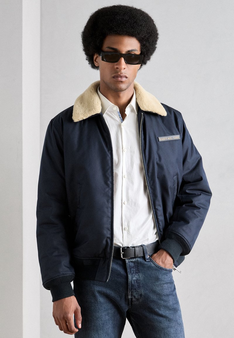 Chaqueta bomber azul marino con un cuello de pelo sintético beige, cierre de cremallera, puños de canalé y un parche con logo. Lleva puesta sobre una camisa blanca y jeans oscuros.