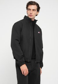 Sort bomberjakke med høj krave, frontlynlås, elastiske manchetter og broderet logo med røde, hvide og blå detaljer.