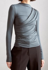 Kvinna klädd i en figursydd, långärmad topp i slategrå med asymmetrisk drapering och svarta högmidjade byxor framför en enfärgad bakgrund.