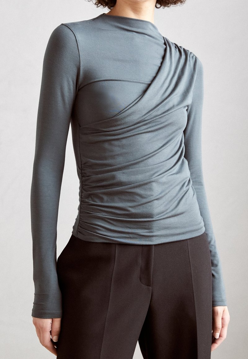 Kvinna klädd i en figursydd, långärmad topp i slategrå med asymmetrisk drapering och svarta högmidjade byxor framför en enfärgad bakgrund.