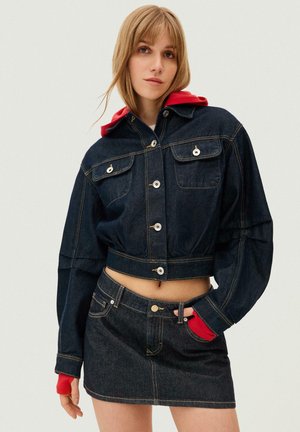 Ung kvinde iført en mørk denim cropped jakke og matchende denim mininederdel med en rød hættetrøje synlig under jakkeærmerne og hætten.