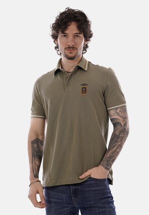 Uomo con capelli ricci che indossa una polo verde oliva con stemma, jeans blu e tatuaggi visibili su entrambi gli avambracci, in piedi davanti a uno sfondo semplice.