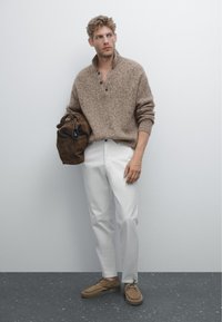 Bruine, gestructureerde gebreide pullover met een kraag, witte broek en bruine suède schoenen. Houdt een bruine leren tas met klepsluiting vast.