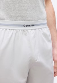 Περιοχή μέσης και άνω γοφών ενός ατόμου που φοράει κοντά σορτς με ελαστική μέση Calvin Klein σε ανοιχτό γκρι και πουκάμισο ανοιχτού γκρι χρώματος.