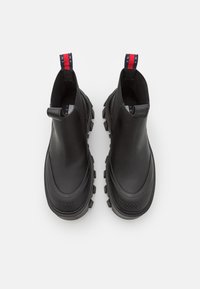 Pár černých středně vysokých gumových bot se silnými podrážkami a textilními poutky se červenými, bílými a modrými pruhy na zadní straně.