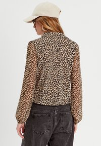 Blusa de leopardo transparente con mangas largas y cuello, combinada con pantalones de mezclilla oscuros de cintura alta. Lleva una gorra beige.