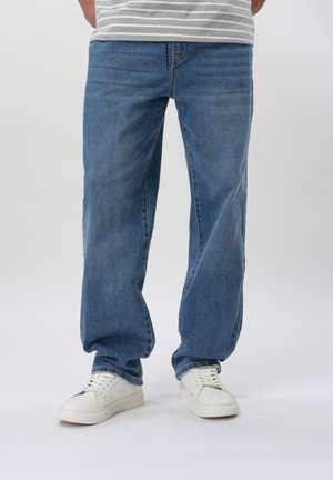 Mann iført blå løstsittende jeans, hvite joggesko og en grå og hvitstripet skjorte, stående mot en ensfarget bakgrunn.