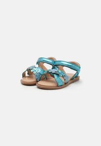 Tyrkise sandaler har blomsterdetaljer, justerbare remmer og en flat såle med en brun bunn, laget av glatte syntetiske materialer.