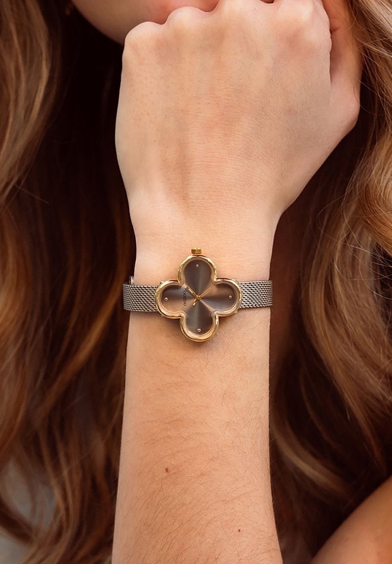 Montre dorée avec un cadran en forme de fleur, bande en maille grise et un cadran translucide présentant des accents dorés et cinq marqueurs horaires.