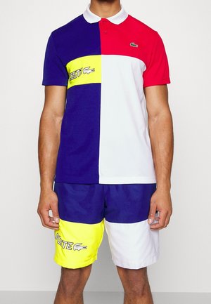 Mężczyzna noszący kolorową koszulkę polo i szorty w kolorach czerwonym, granatowym, białym i żółtym z logo Lacoste na koszulce i szortach.