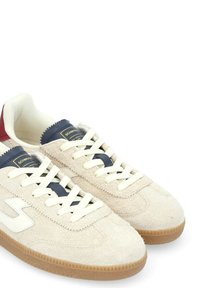 Sneakers in suede beige con lacci bianchi e una linguetta blu navy, dotati di una suola in gomma e un accento logo bianco di lato. Dettaglio del tallone rosso.