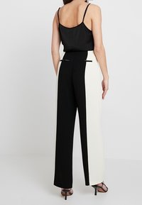 Kvinna som står vänd bort, klädd i en svart utan ärmar topp och högmidjade vida byxor med svarta och vita vertikala paneler.