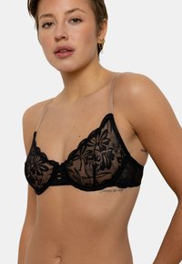 Soutien-gorge en dentelle noire avec un motif floral, des bonnets triangulaires et des bretelles transparentes. Il est orné d'un accent central en métal doré et de bords festonnés.