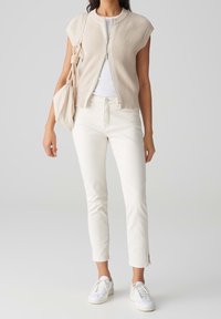 Ljusbeige stickad väst med korta ärmar, vit t-shirt under, smala vita byxor och vita sneakers. Väska i matchande färg.