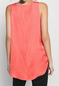 Korallfärgad ärmlös topp med rundad nederkant, slät textur och rynkad halsringning, ihopparad med svarta byxor för kontrast.