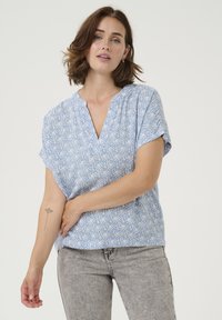 Haut court à manches courtes bleu clair avec un décolleté en V, présentant un motif abstrait blanc. Le tissu semble doux et léger, idéal pour une tenue décontractée.
