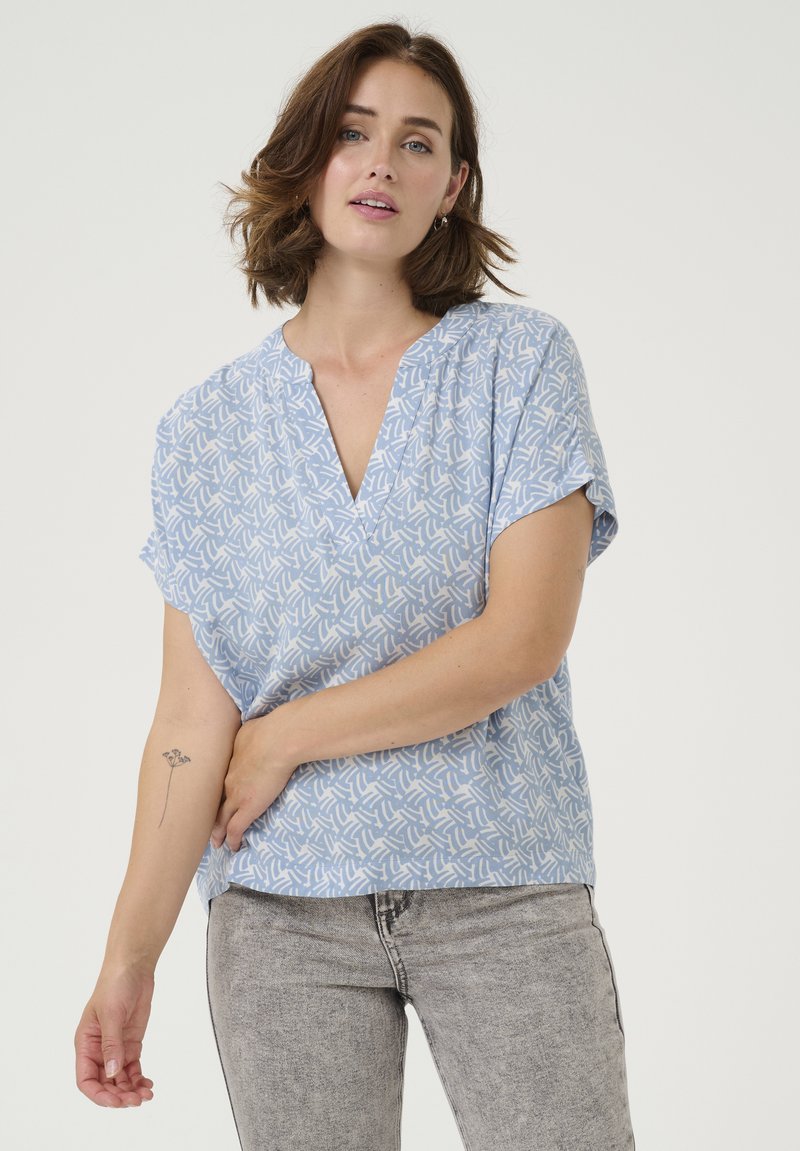 Haut court à manches courtes bleu clair avec un décolleté en V, présentant un motif abstrait blanc. Le tissu semble doux et léger, idéal pour une tenue décontractée.