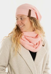 Roze gebreide beanie met een slouchy ontwerp en leren label, gecombineerd met een bijpassende roze infinity sjaal met een gestructureerd patroon.