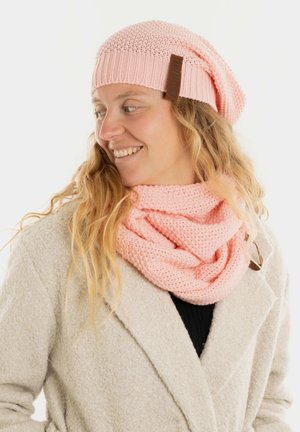 Roze gebreide beanie met een slouchy ontwerp en leren label, gecombineerd met een bijpassende roze infinity sjaal met een gestructureerd patroon.