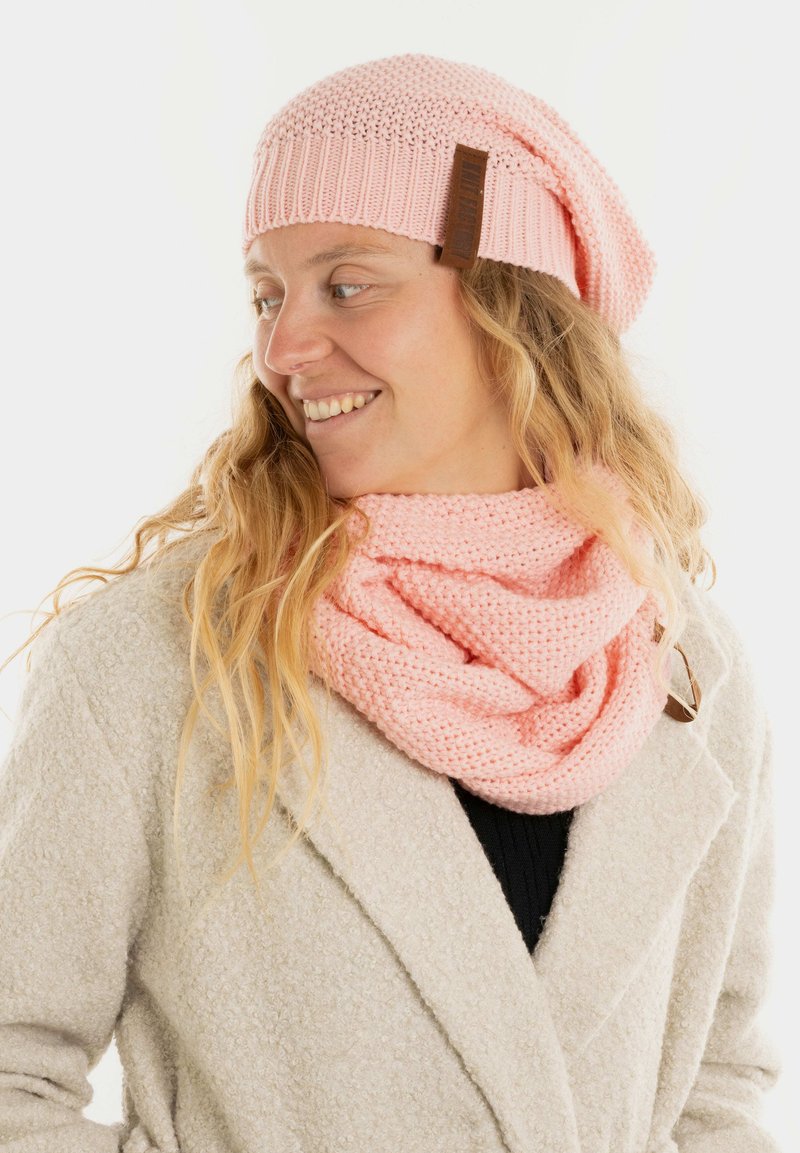 Roze gebreide beanie met een slouchy ontwerp en leren label, gecombineerd met een bijpassende roze infinity sjaal met een gestructureerd patroon.