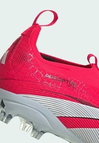 Scarpino da calcio rosso con tomaia sintetica testurizzata, linguetta laterale, tacchetti traslucidi e dettagli argento sulla suola.