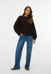 Frau mit langen Haaren, die einen dunklen, übergroßen Pullover, blaue weite Jeans und braune Schuhe trägt, steht mit einer Hand in der Tasche.