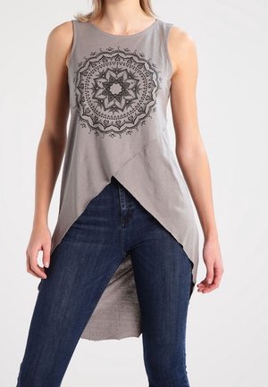 Femme portant un haut gris sans manches avec un motif mandala noir et un ourlet asymétrique sur un jean bleu foncé.
