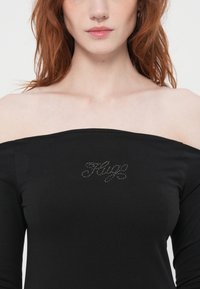 Svart off-shoulder topp laget av mykt stoff med jevn tekstur, med "Hug" i sølv rhinstein i midten.