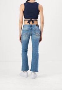 Jeans de mezclilla acampanados en azul claro con una textura desgastada, que presentan bolsillos traseros y un parche con el logo en la cintura. Combinados con un top corto azul marino.
