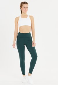 Sujetador deportivo blanco y mallas de cintura alta en verde oscuro con bolsillos laterales. Las mallas tienen una textura suave y una forma ceñida.