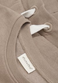 Vêtement en coton beige avec un col et des poignets côtelés, présentant des liens en tissu blanc et une étiquette inscrite "minymo" à l'intérieur.