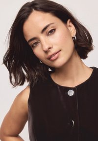 Mustast värvist varrukateta rõivas ümarate kaelustega ja läikivate nööpidega. Modelil on õlgadeni ulatuvad lainelised tumedad juuksed ja neutraalne meik.