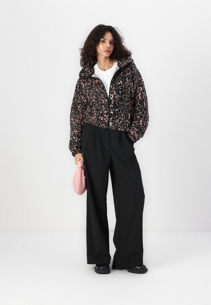 Giacca cropped con stampa leopardata nera e rosa, con zip frontale e cappuccio, abbinata a pantaloni neri a gamba larga e una borsa rosa.