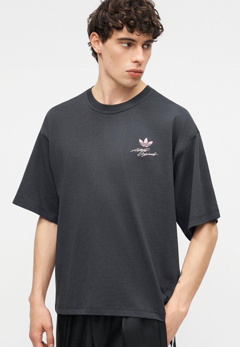 Young man with curly hair wearing a loose black Adidas t-shirt with pink logo and signature, looking to his left, hand in pocket.