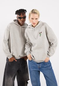 Graue Hoodies mit Fronttaschen, die ein farbenfrohes V-Logo zeigen; das Model kombiniert sie mit lockeren Jeans, einer trägt Sonnenbrillen.