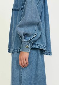 Gai zila džinsu krekls ar garām, apjomīgām piedurknēm, ar pogājamām aprocēm un brīvu piegriezumu. Iezīmē kontrastējoša šuvuma un krūšu kabatu.