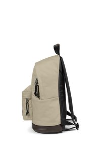 Béžový látkový batoh s černými stranami a dnem. Obsahuje nastavitelné popruhy, vnější kapsu se stahovací šňůrkou a horní poutko pro uchopení.