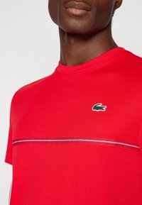 T-shirt di cotone rossa con collo a girocollo, caratterizzata da un logo di coccodrillo verde e un design a righe orizzontali sul petto in colori a contrasto.